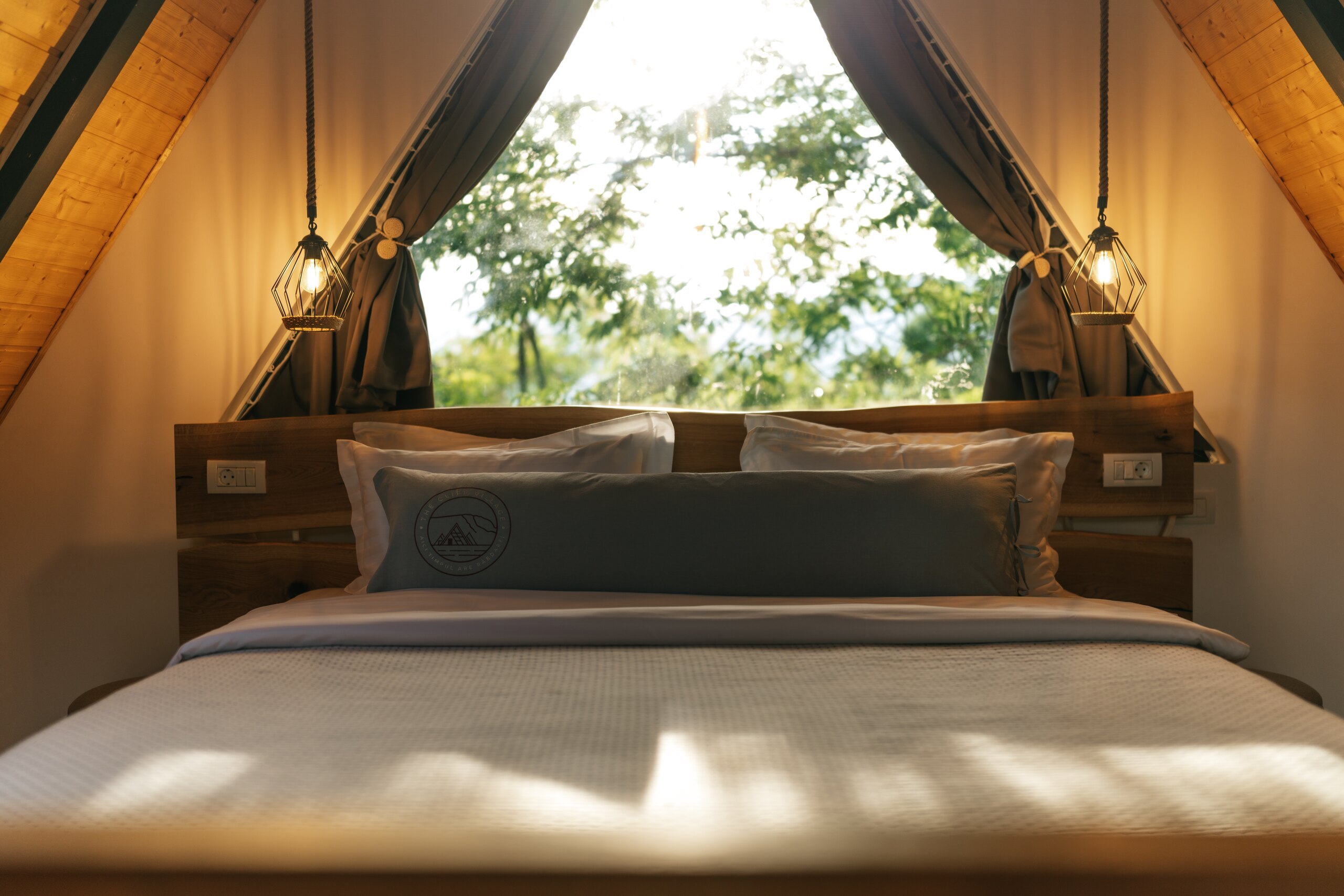 A made bed under the A-frame roof, two lamps and a window onto the trees