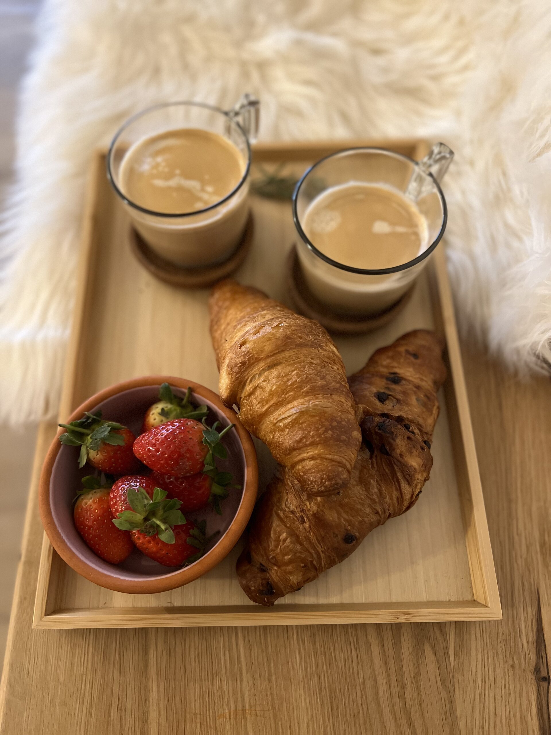 A wooden tray with two coffees, a croissant and a bowl of strawberries on a sheepskin