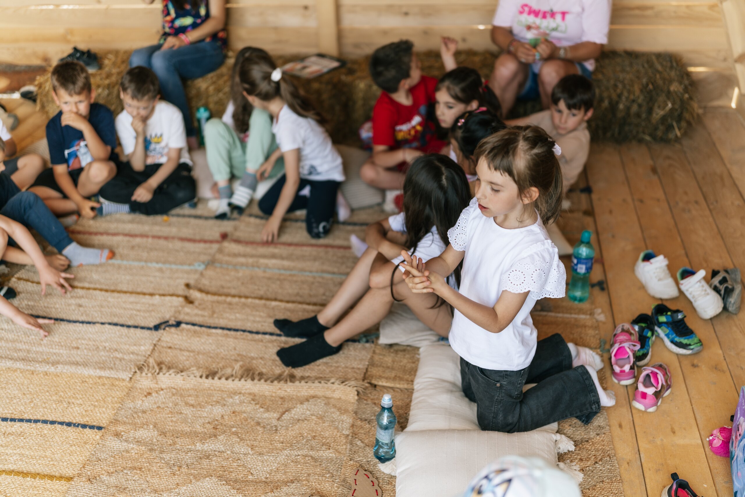Children sit on rugs and straw bales in a wooden shelter, listening and laughing