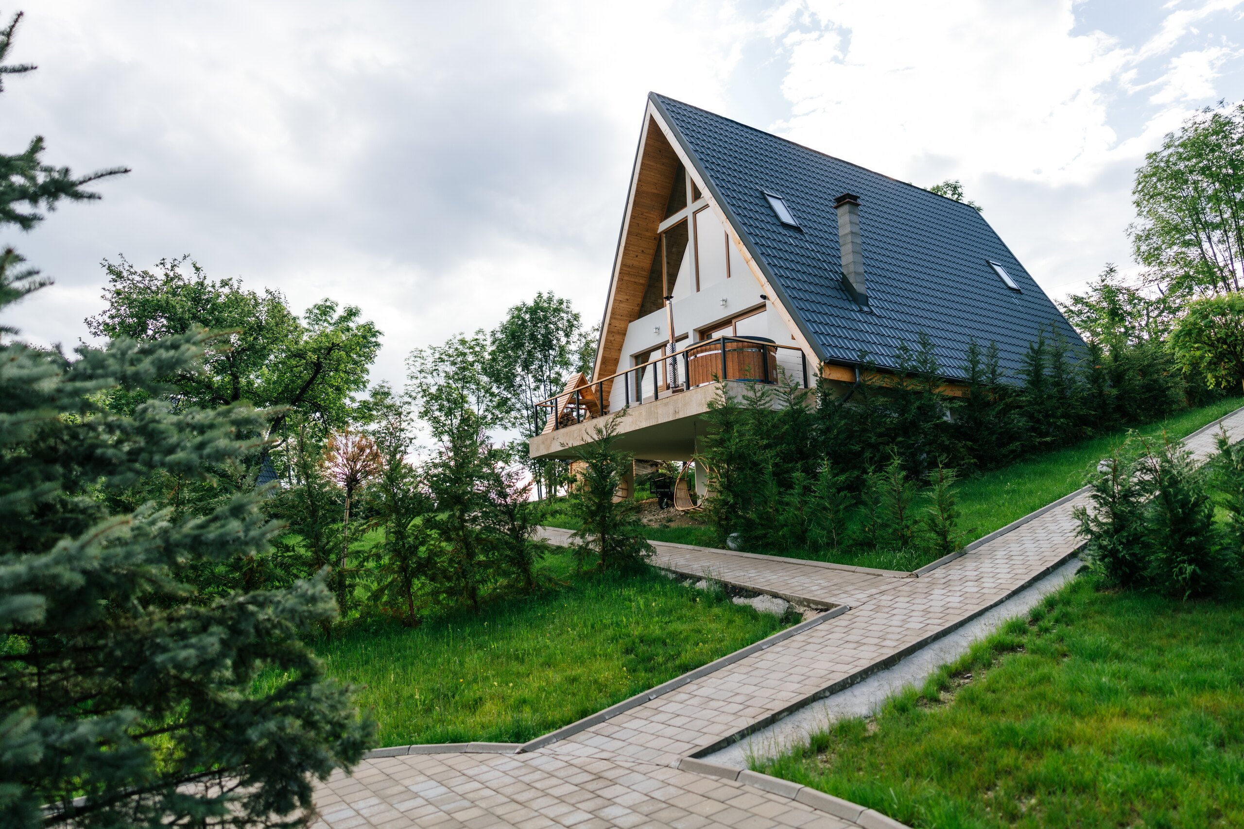 A dark A-frame chalet with a terrace above a lawn, a stone path leading up to it