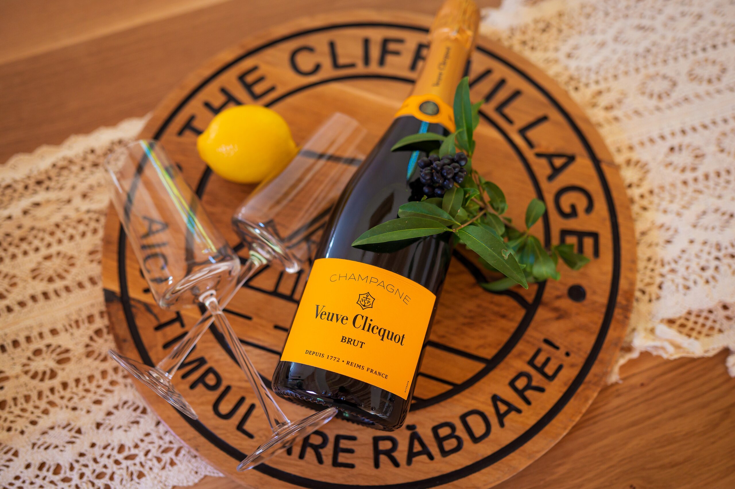 A bottle of champagne, a glass and a lemon on a round wooden board with the village name