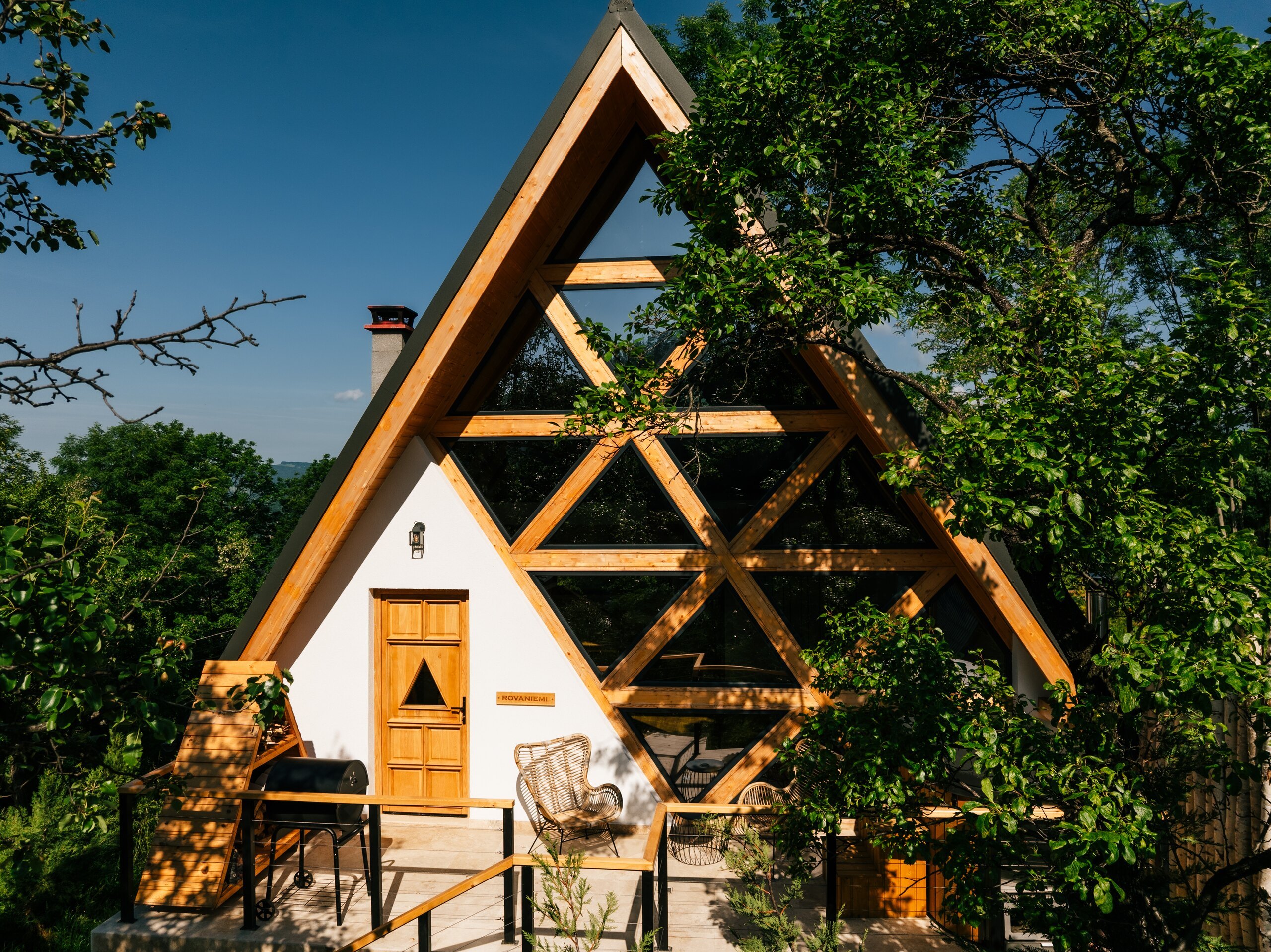 The front of a white A-frame cottage with a wooden lattice, a small terrace and a tree beside it