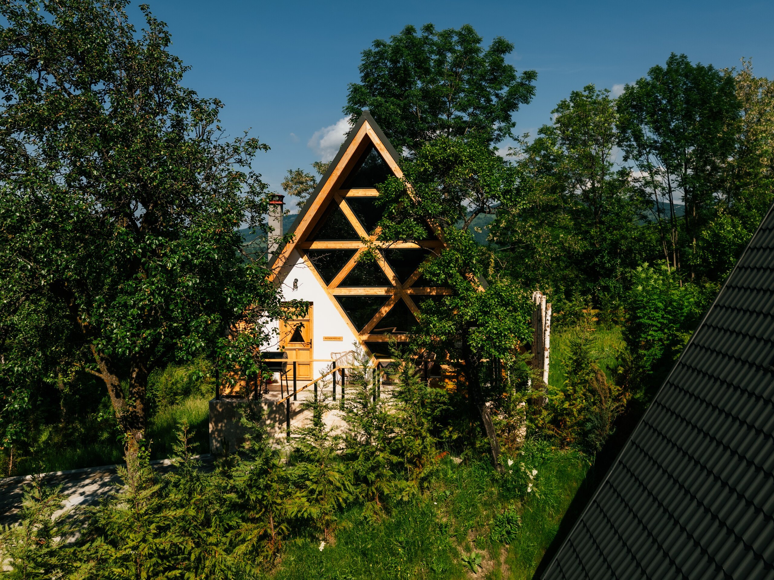 A small A-frame cottage with a wooden lattice front, half hidden by summer trees