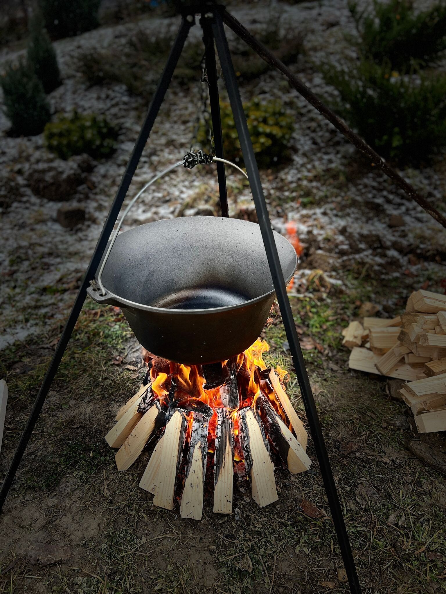 A black cauldron hangs from a tripod over an open fire at dusk