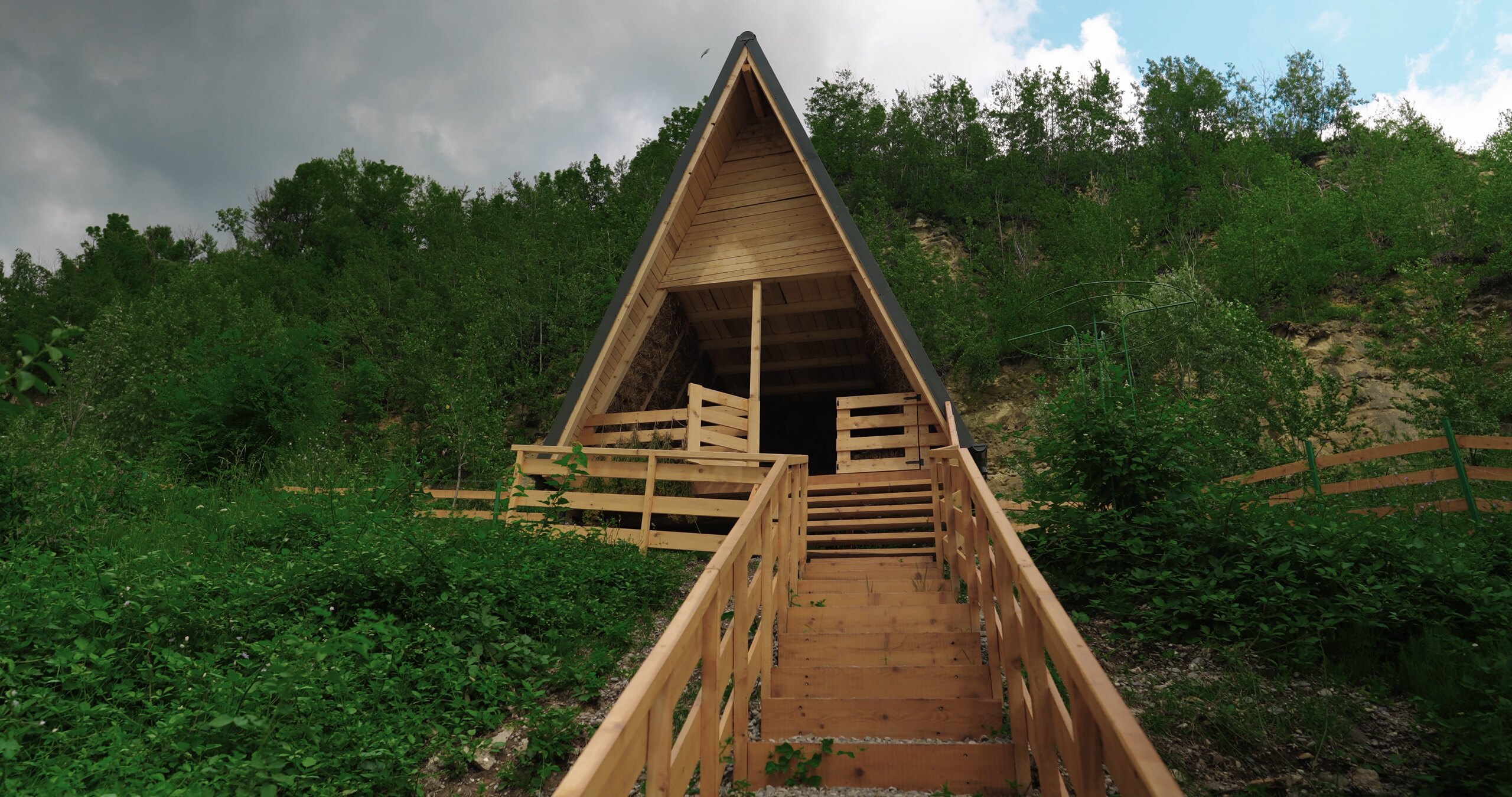 A small wooden A-frame hut with a railing on a hillside, trees behind