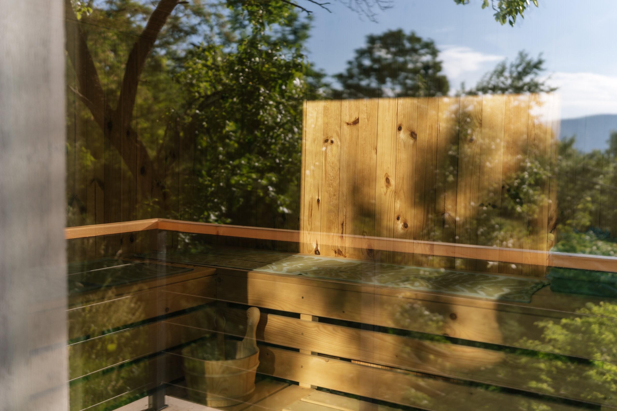 A wooden hot tub on a terrace, trees and sky reflected in the glass