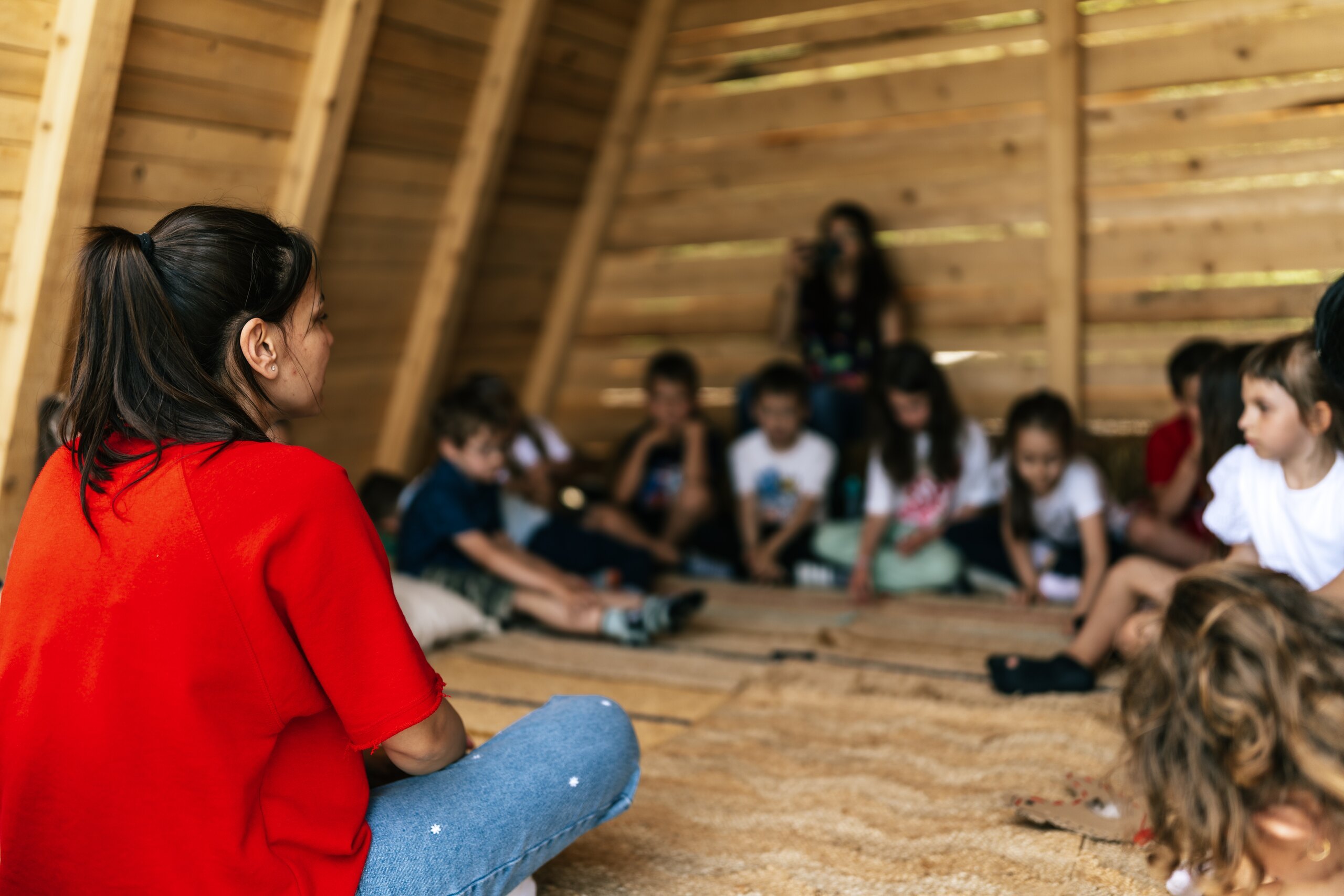 A woman in red talks to a circle of children sitting on rugs in a wooden shelter