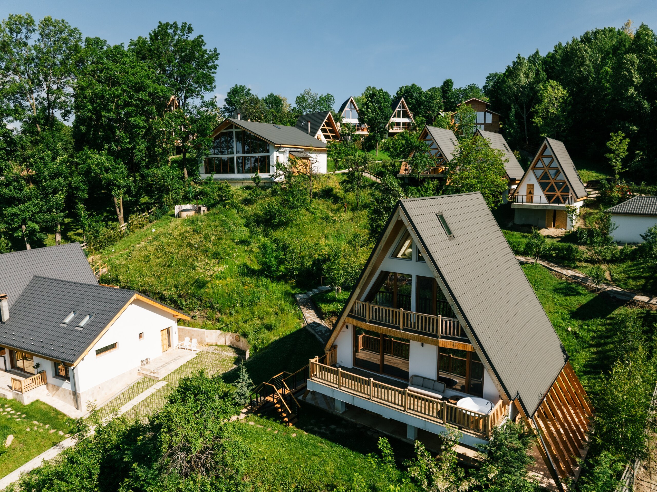Three A-frame houses with wooden terraces on a grassy slope, trees behind