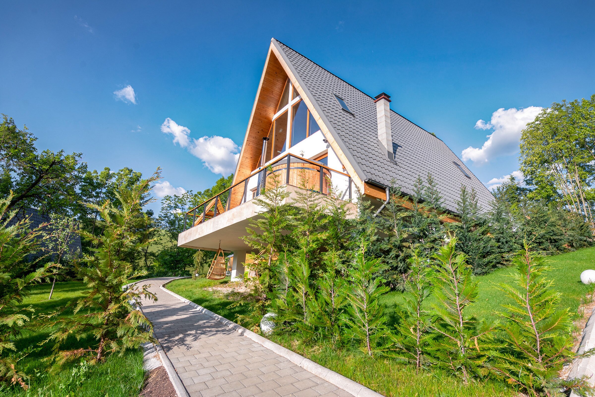 A large A-frame villa with a balcony, a stone path and young trees in front